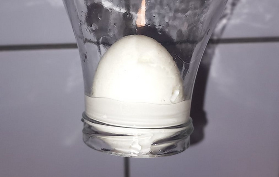Nahaufnahme von einem hartgekochten Ei, das sich in einer Glasflasche befindet. Die Glasflasche wird verkehrt herumgehalten und das Ei drückt von innen auf die Flaschenöffnung